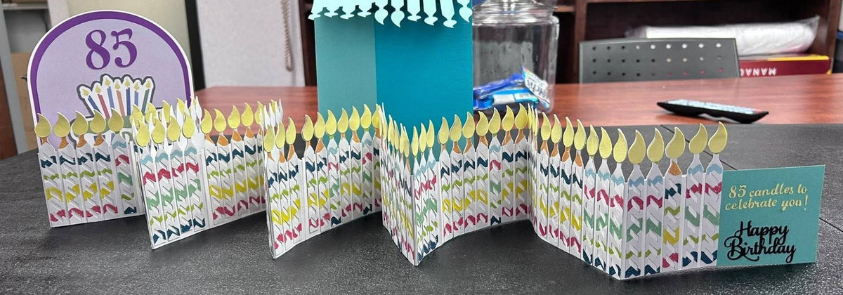 Handmade accordion birthday card extended to show a long row of layered white paper candles with colorful stripes and yellow flames, an 85 arch panel, and a teal Happy Birthday end panel