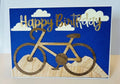 Handmade birthday card with a layered wood-grain bicycle on wood-grain hills, cream paper clouds, and gold-foil Happy Birthday lettering on a blue textured background