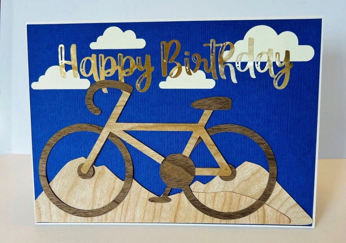 Handmade birthday card with a layered wood-grain bicycle on wood-grain hills, cream paper clouds, and gold-foil Happy Birthday lettering on a blue textured background