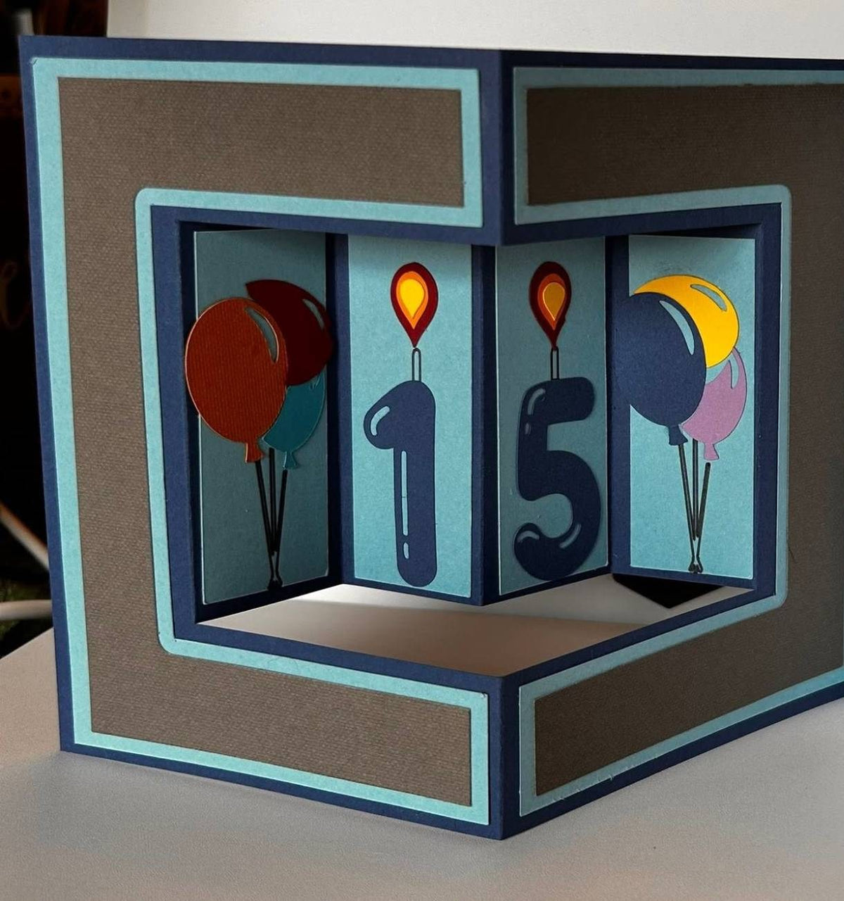 Handmade 3D box-style birthday card opened to a layered navy, light-blue, and brown scene with colorful paper balloons and candle-style number digits