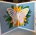 Handmade pop-up card opened to show a white cut-paper butterfly with watercolor-style wings standing over a layered yellow paper flower and green leaves on light-blue pages