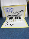Handmade pop-up card opened to show raised paper piano keys, a curling keyboard ribbon, black music notes, and Wishing you the best in blue script on a cream and yellow card
