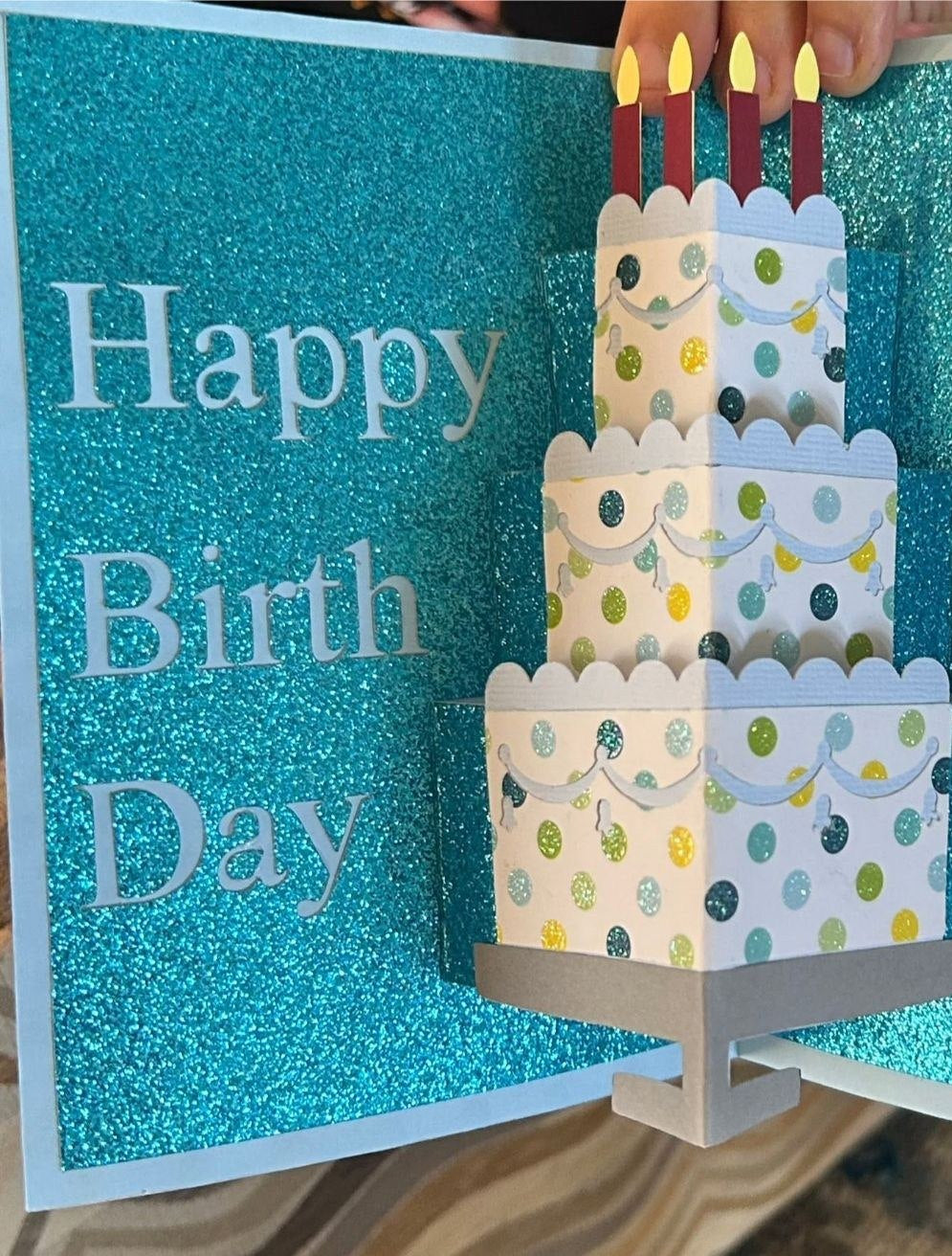 Handmade pop-up birthday card with teal glitter pages, a three-tier white polka-dot paper cake with four candles on a gray stand, and Happy Birth Day lettering