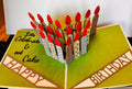 Handmade pop-up birthday card with green pages, a cluster of patterned paper candles with red flames, Happy and Birthday banners, and Let’s Celebrate and eat Cake in script