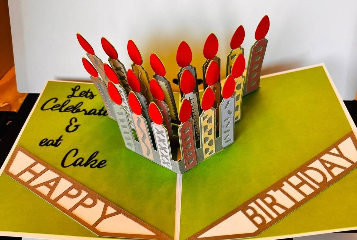 Handmade pop-up birthday card with green pages, a cluster of patterned paper candles with red flames, Happy and Birthday banners, and Let’s Celebrate and eat Cake in script
