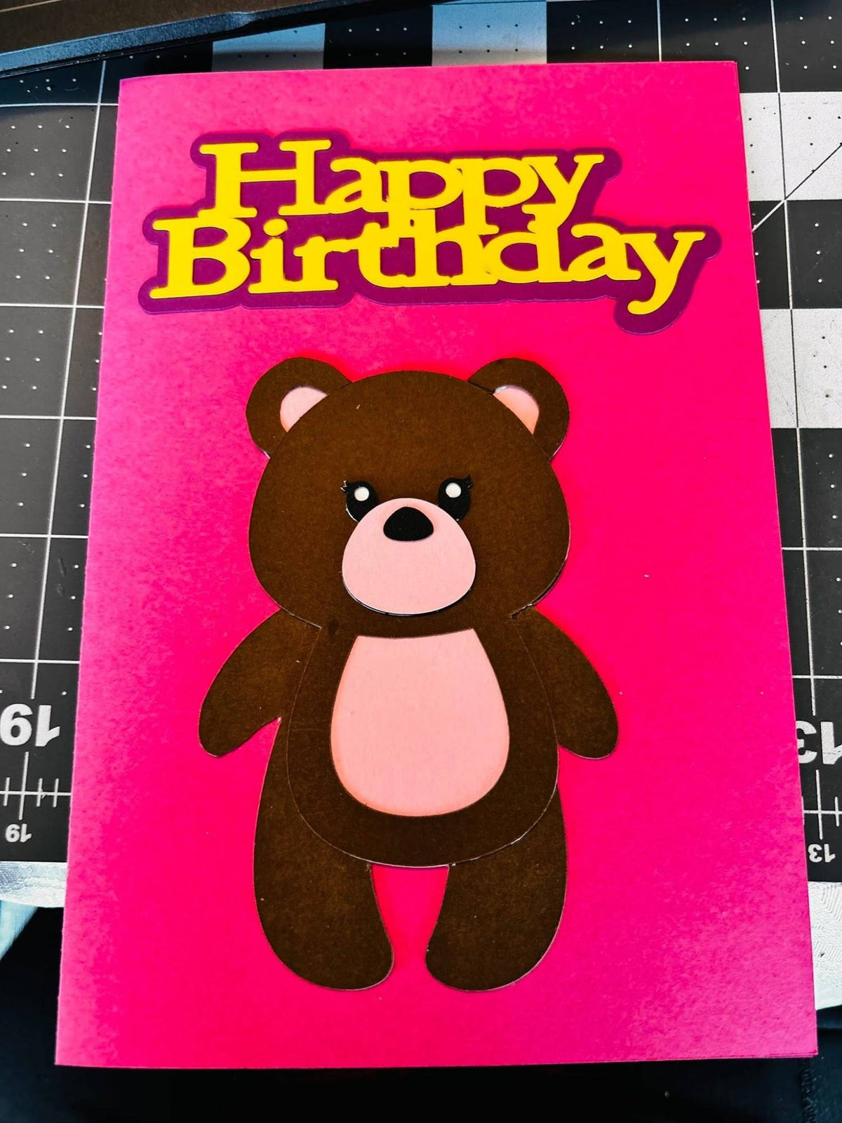 Handmade hot-pink birthday card with a layered brown paper teddy bear with pink ears and tummy under yellow Happy Birthday lettering with a purple shadow