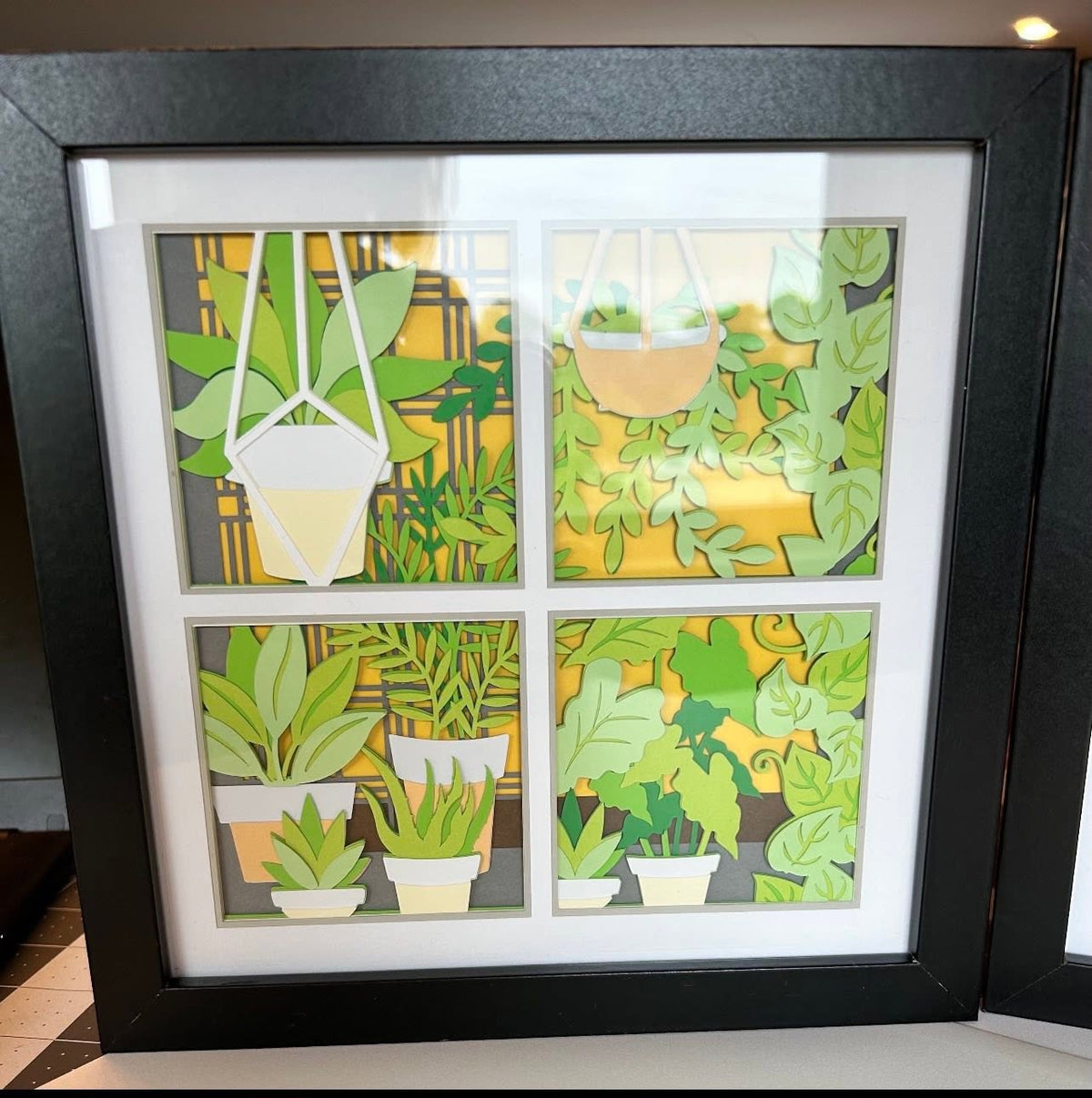 Handmade four-pane plant window layered paper shadow box with hanging planters and potted plants in a black frame